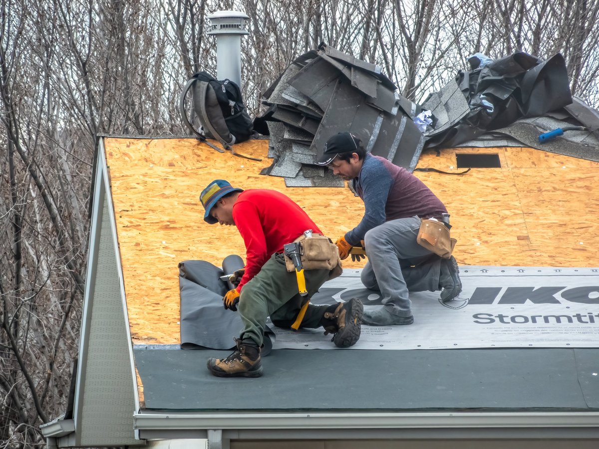 Two construction workers installing black underlayment on a plywood roof, preparing for shingles, tools and bundles of shingles visible